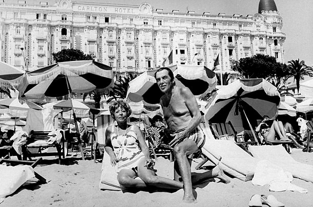 TV emcee Ed Sullivan kneeling beside wife Sylvia on beach w. other sunbathers behind them & Carlton Hotel in bkgrd., during vacation.