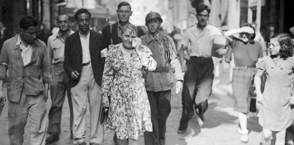 A sobbing French woman with a swastika smeared on her face is paraded through the streets with civilians and a soldier.