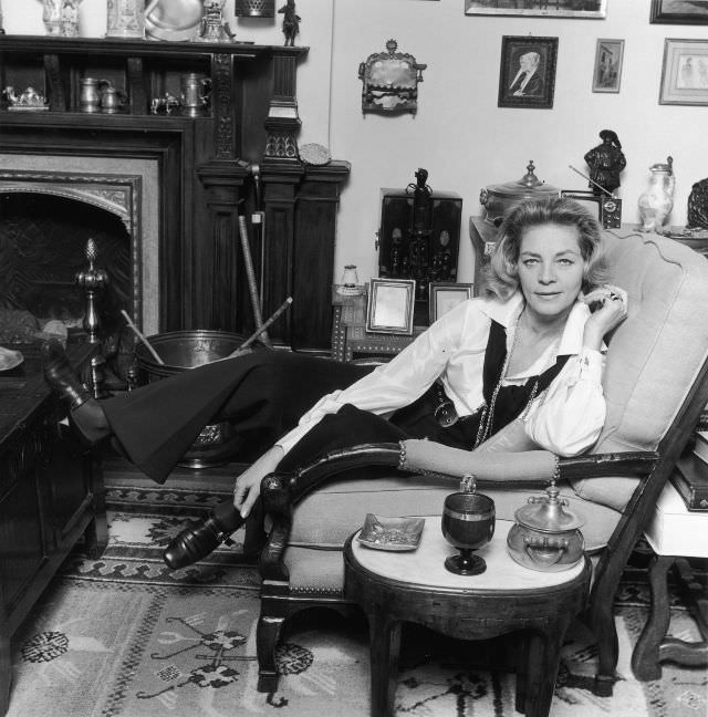 Lauren Bacall sitting in an armchair with one leg propped on a table, at her home, March 1969.