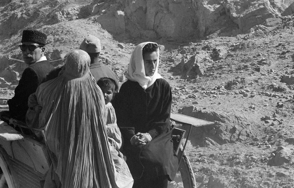 Afghan women, men, and child in traditional dress ride in a cart through an arid, rocky landscape, November, 1959.