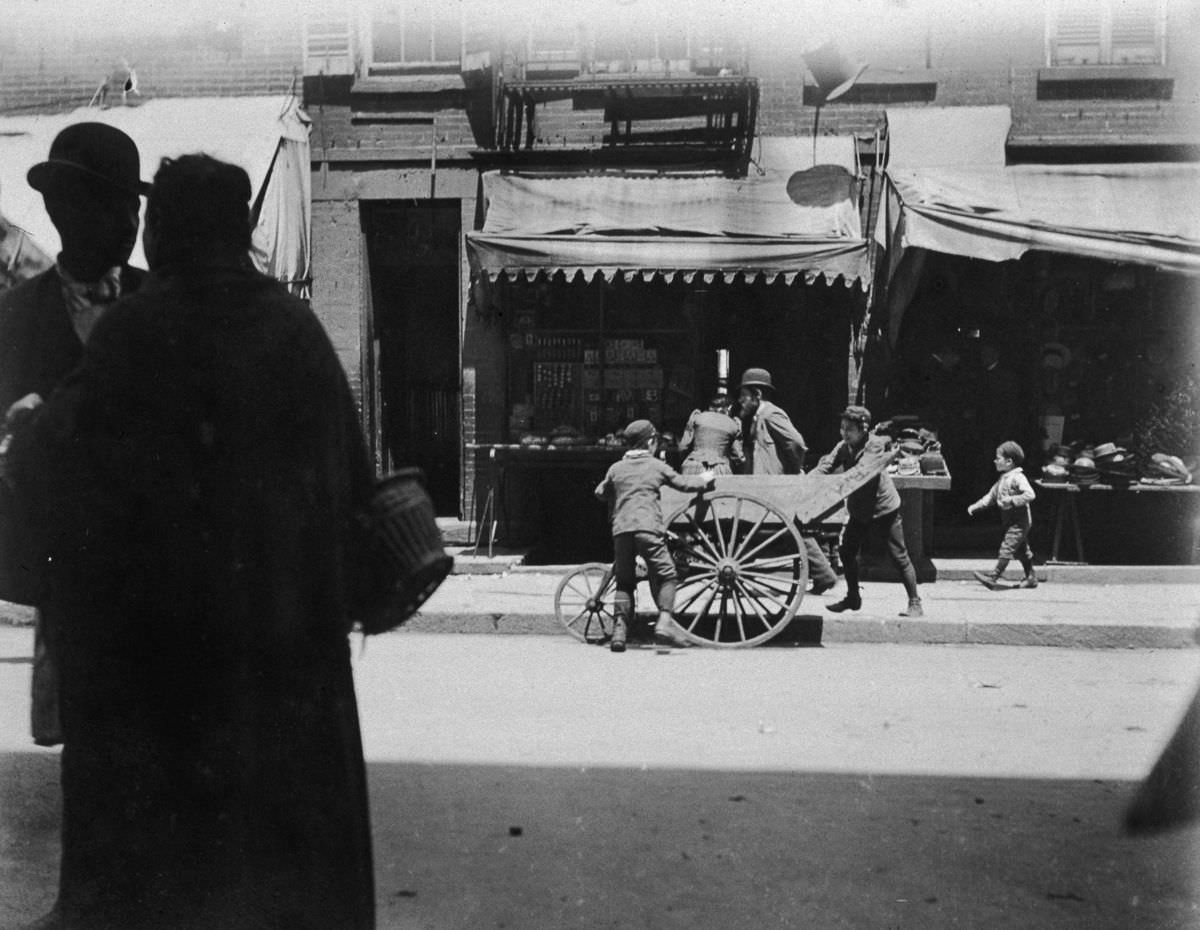 Two young boys laugh and steal items from a vendor's pushcart on Hester Street in the Lower East Side, 1895