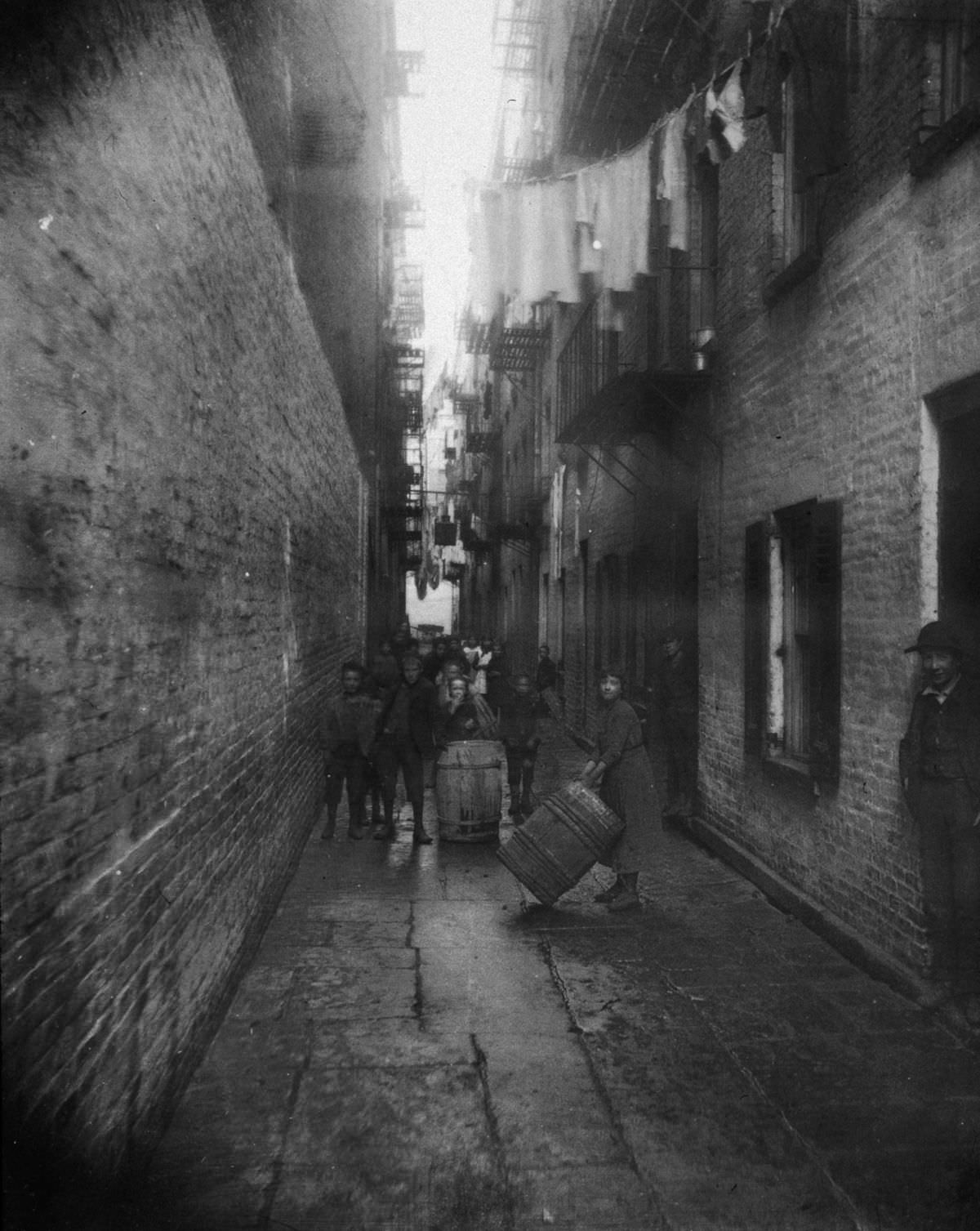Children play with barrels in an alley between tenement buildings in Gotham Court, 38 Cherry Street, 1890