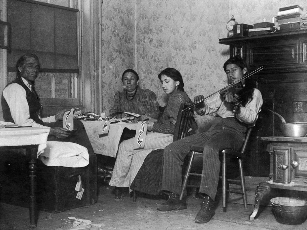 Mountain Eagle and his family make Native American handicrafts while his son plays violin in their tenement at 6 Beach Street, 1895
