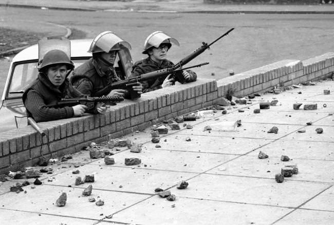 Three British soldiers, two armed with automatic rifles, and man at left with a Stirling sub-machinegun, shelter behind a wall in the Andersonstown area of Belfast, Northern Ireland on Nov. 1, 1971, during riots which followed the shooting of two policemen.