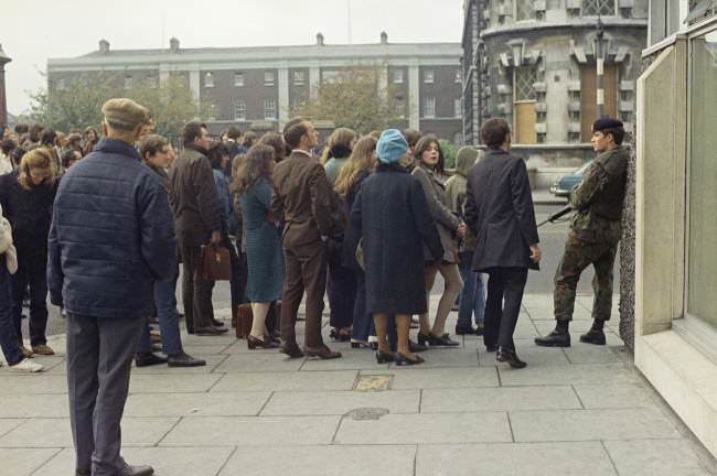 A British soldier stands guard as bystanders wait to get a view of operations by the army bomb disposal squad in Northern Ireland on Nov. 11, 1971 after an explosive device had been planted near the city centre.