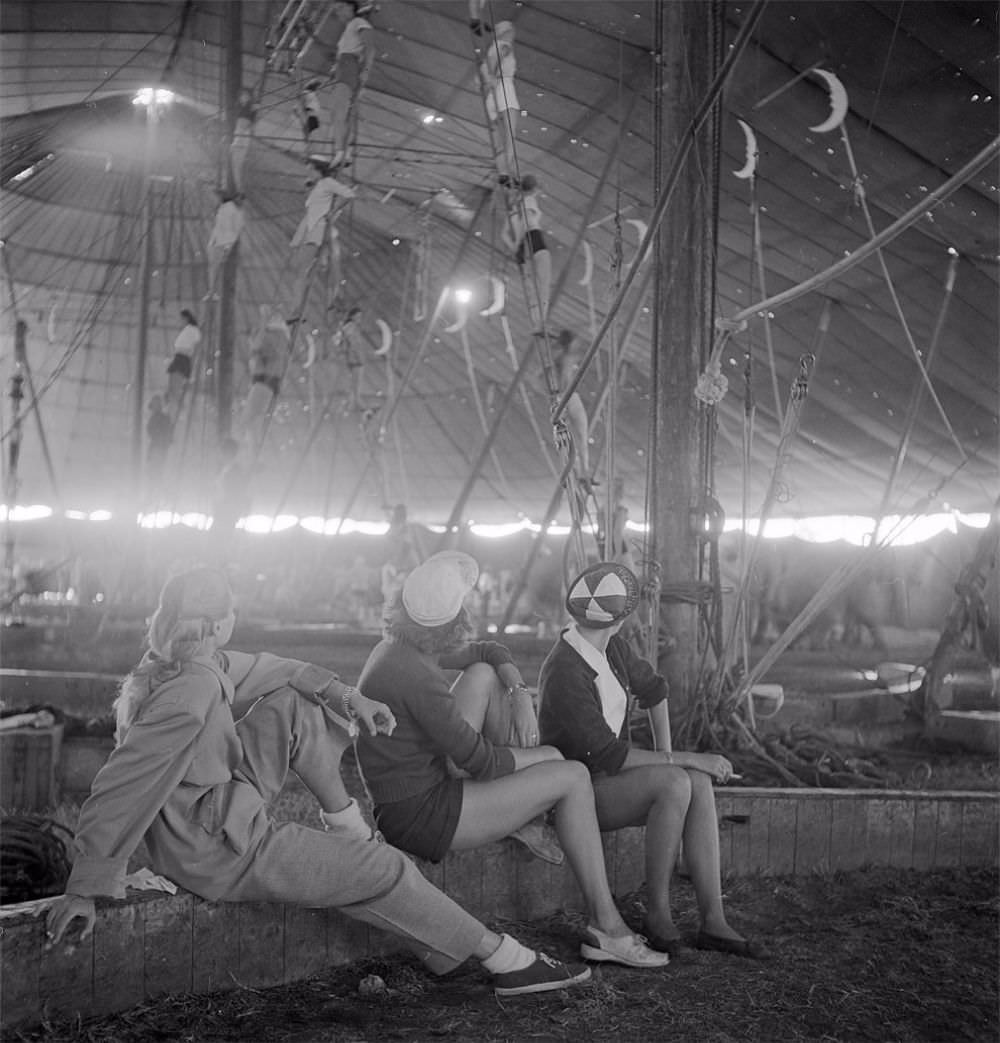 Three circus girls sitting on a wooden plank and watching a group of aerialists rehearsing with ropes for the Ringling Bros. and Barnum & Bailey Circus in Sarasota, FL in 1949.