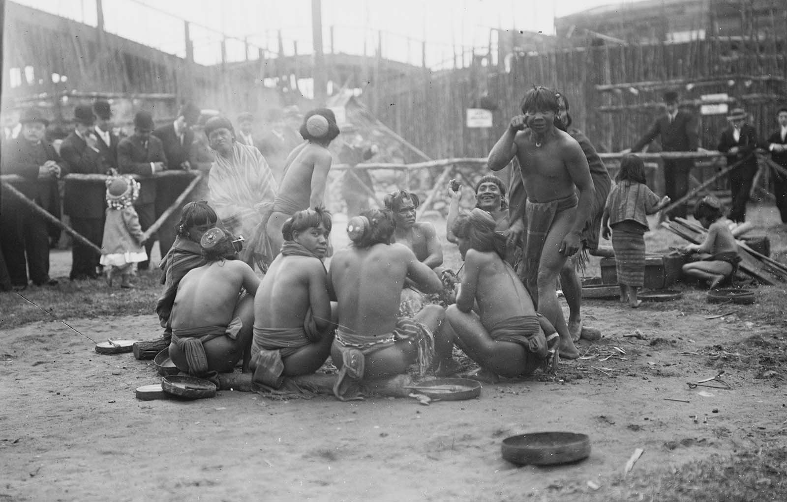 Filipinos in loin cloths sitting in a circle together at Coney Island in New York in the early 20th century while crowds of Americans watch on from behind barriers.