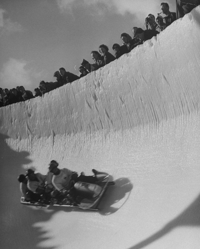Good of cresta, a kind of bobsled, coasting around sunny bend as people peer from above the cresta run.