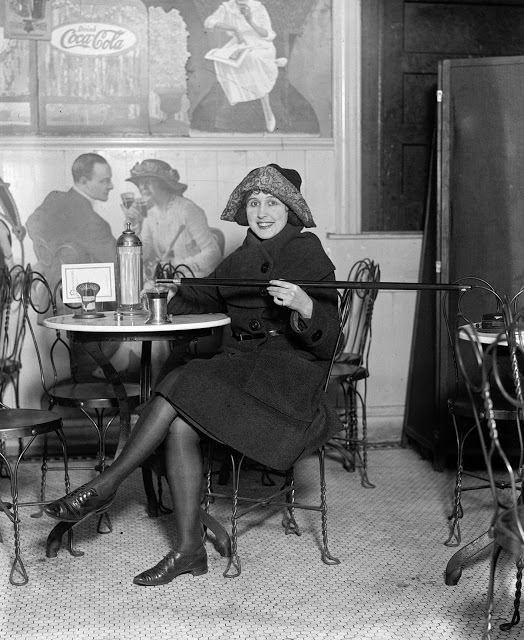 Woman seated at a soda fountain table pours alcohol into her cup from a cane. Note the large Coca-Cola advertisement on the wall, 2/13/1922.