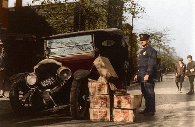 Policeman stands alongside a wrecked car and several cases of moonshine.