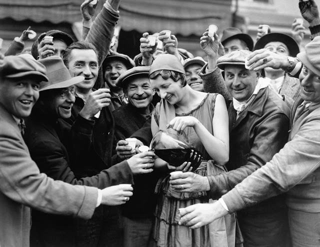 A woman serves drinks to a crowd of men who are joyfully celebrating the repeal of Prohibition in 1933.
