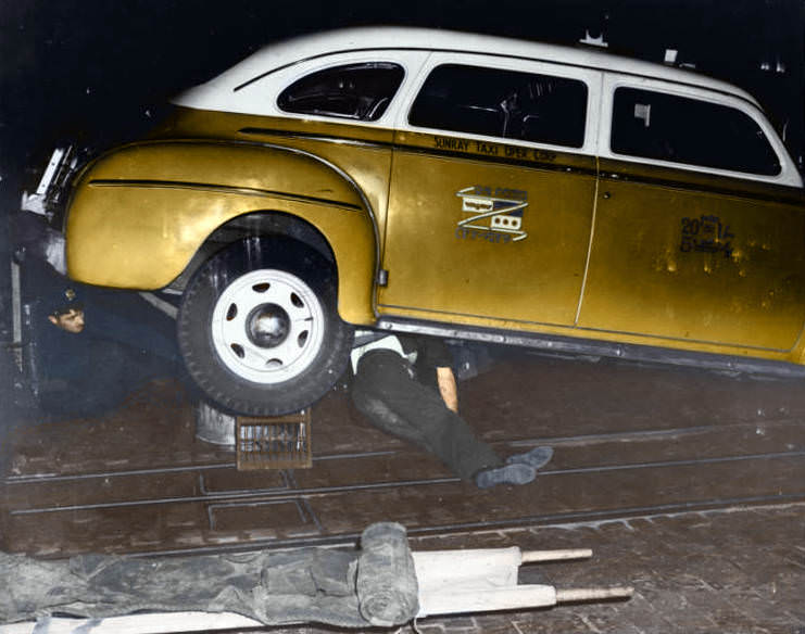 A police officer crouches under the rear end of a taxi jacked up on a crate and garbage can as the dead body of a man who was hit by the cab lies underneath, 1943.