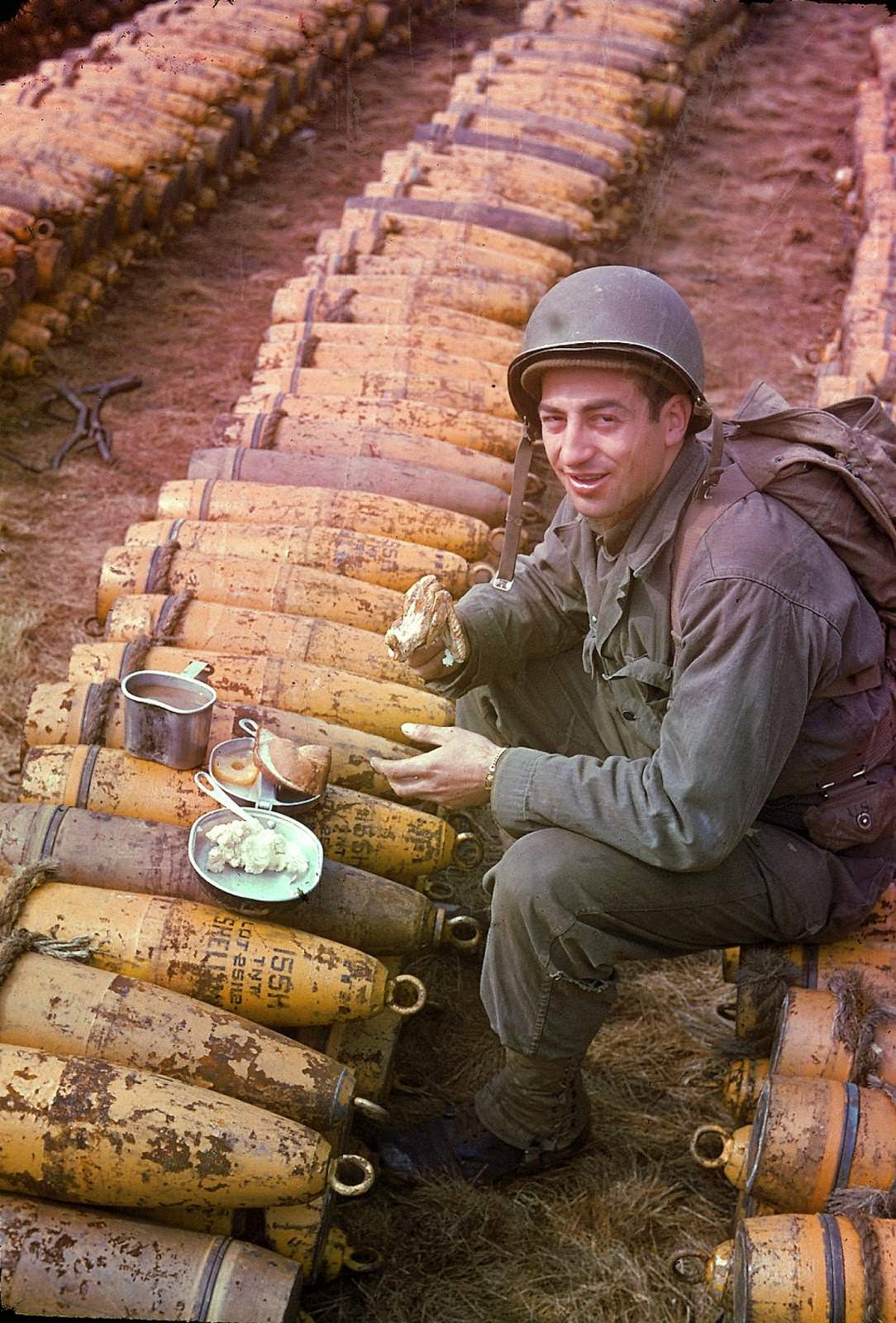 American soldier sits and eats his meal (which includes chicken, mashed potatoes, bread, and pineapple) atop rows of a stockpiled ammunition shells, England, May 1944.