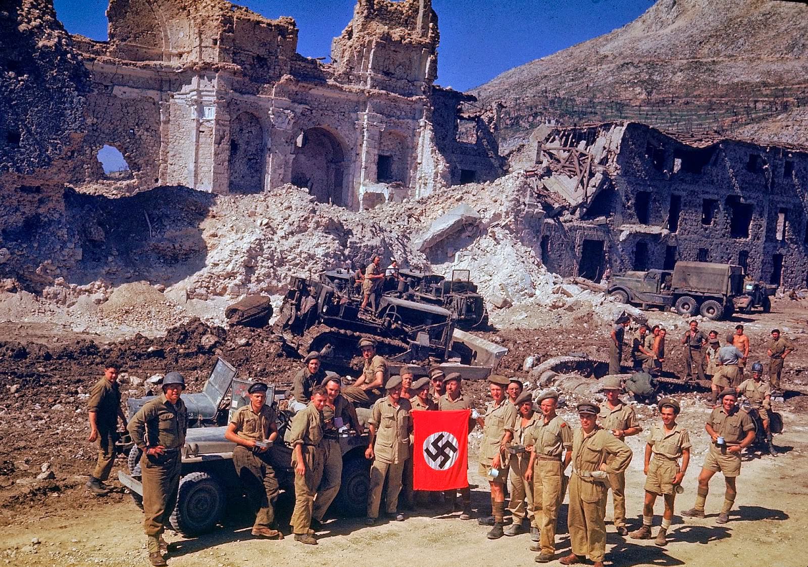 British and South African soldiers hold up Nazi trophy flag while combat engineers on bulldozers clear a path through the debris of the bombed-out city of Cassino in 1943.