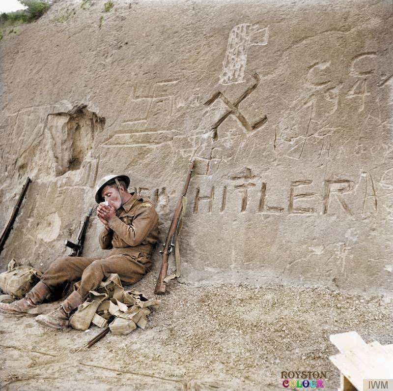 Allied Soldier takes a break during the approach to Tripoli, Libya beside a swastika and the words 'Heil Hitler' that have been carved into a rocky hillside during January 1943