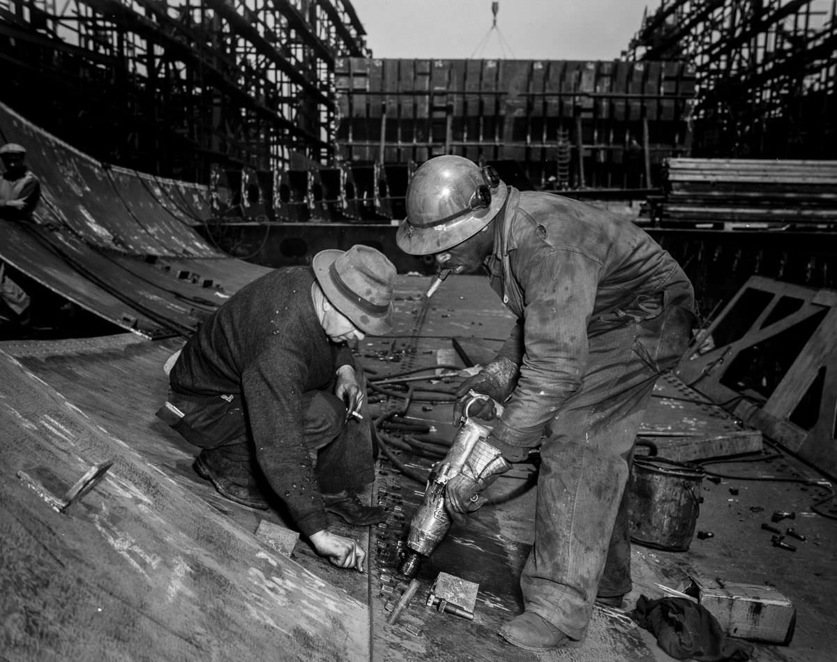 Workers tighten bolts with a pneumatic wrench in the belly of the Frederick Douglass