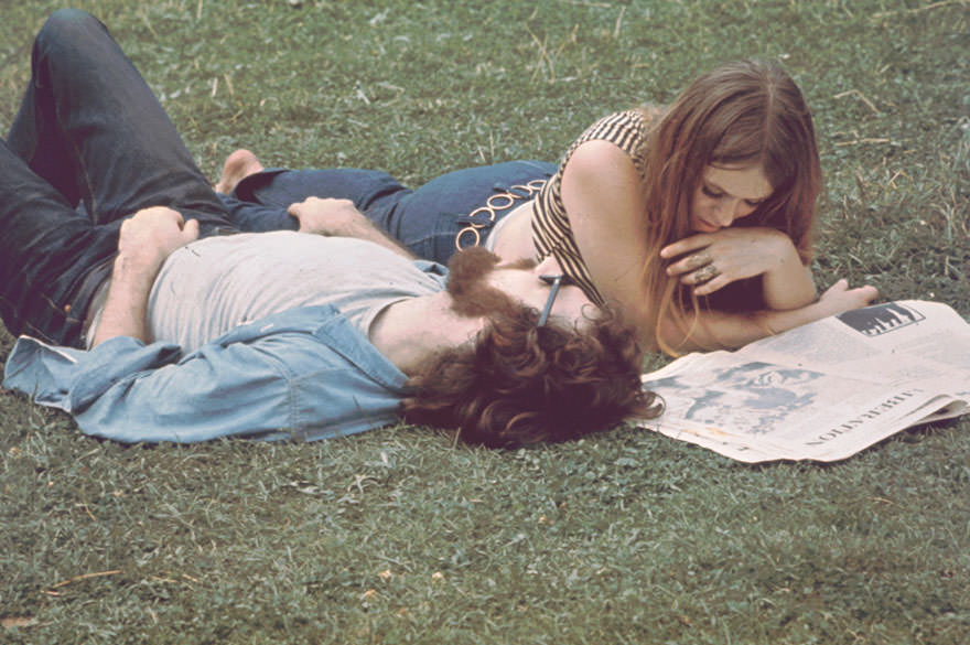 A Young Woman Lies On Grass And Reads Newspaper, A Young Man Beside Her, During The Woodstock Music And Arts Fair