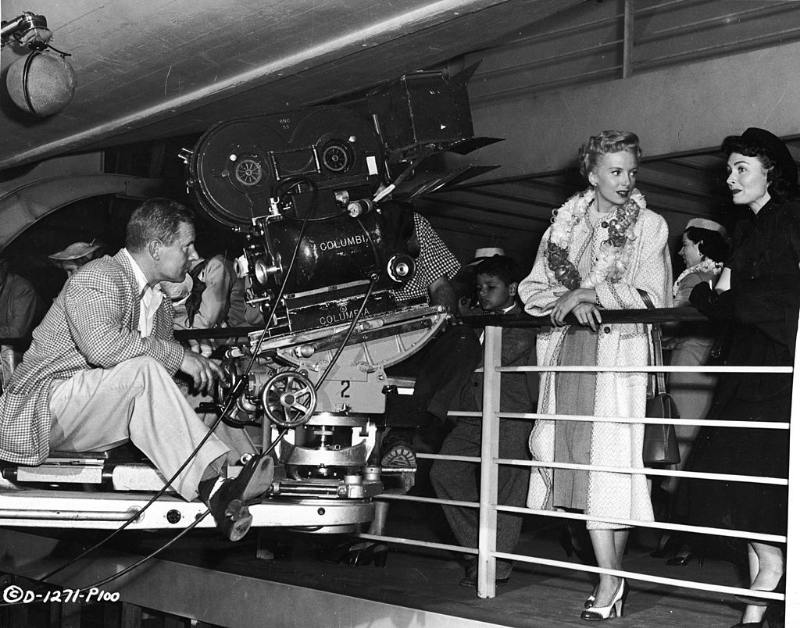Deborah Kerr and Donna Reed standing by a railing while cameraman Kit Carson rolls film on the set of 'From Here To Eternity,' 1953