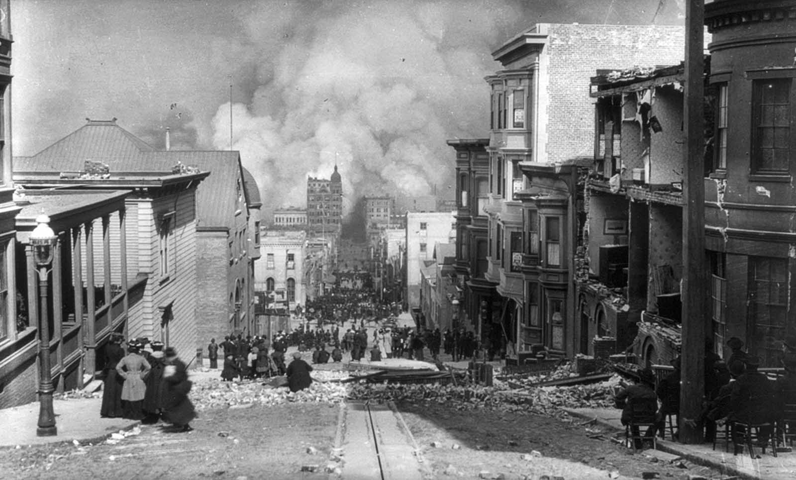 San Francisco residents, some seated in chairs, sit among the earthquake damage, watching out-of-control fires in the distance