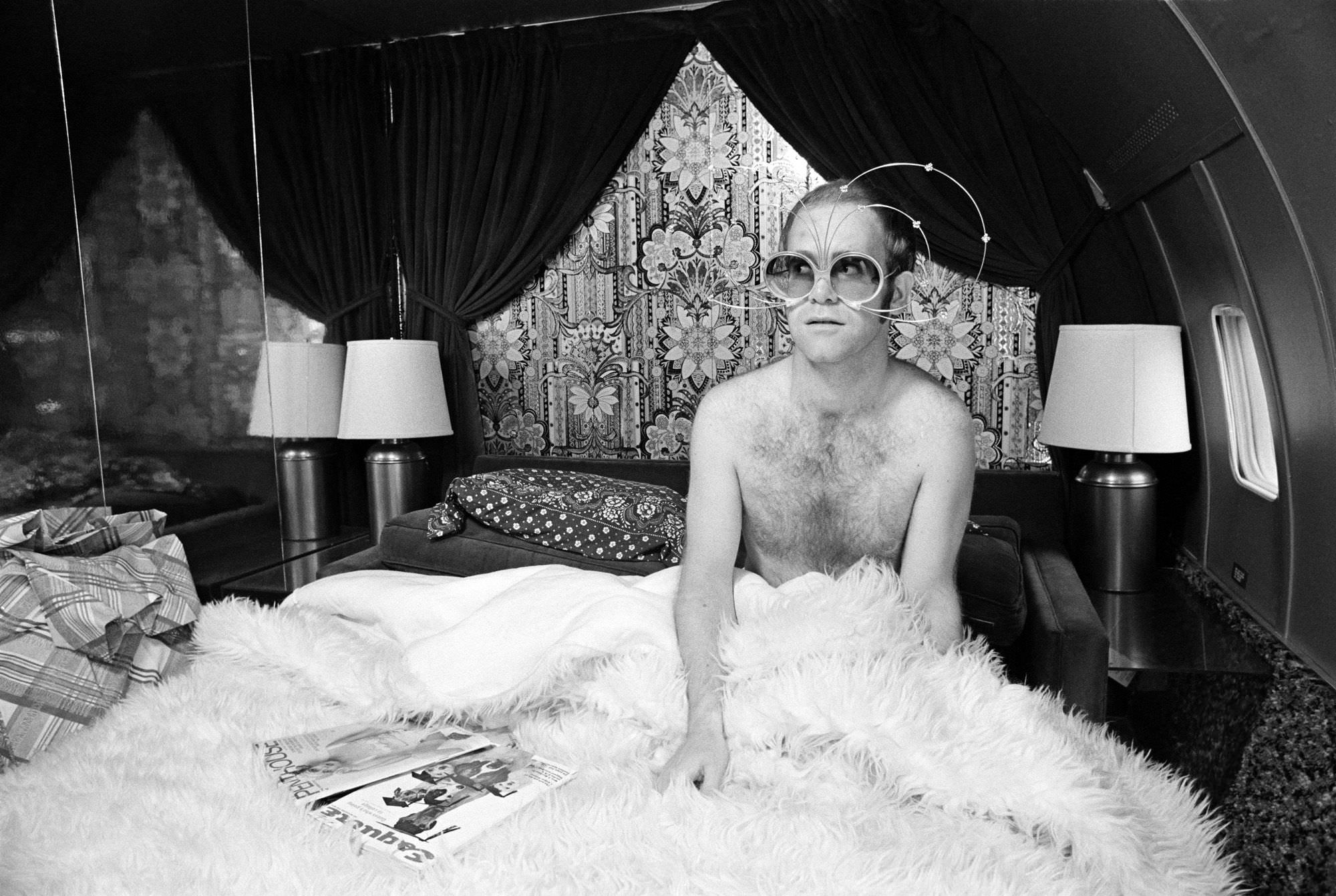 Elton John photographed by Terry O’Neill in bed on a private plane, 1975.