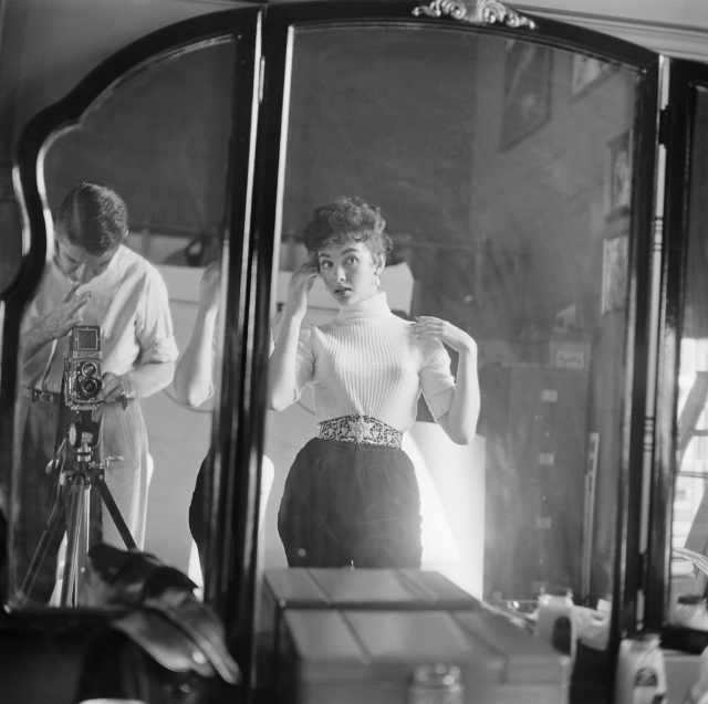Rita Moreno checking her hair and makeup in the mirror while the photographer prepares to take her photograph, Hollywood, 1954.