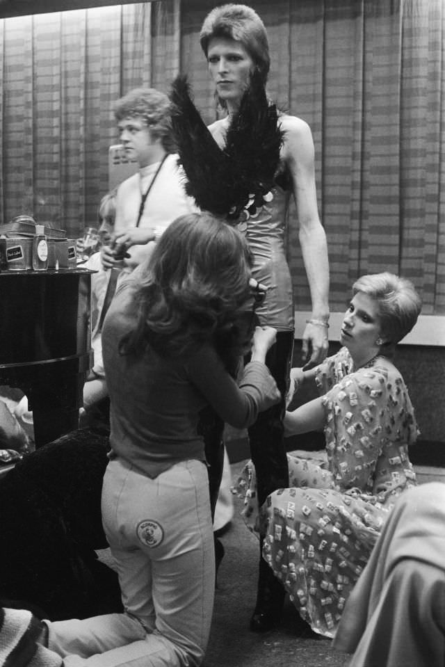 David Bowie, preparing for a performance backstage, circa 1973. He is being dressed in an extravagant costume by his wife Angie Bowie (right) and an assistant