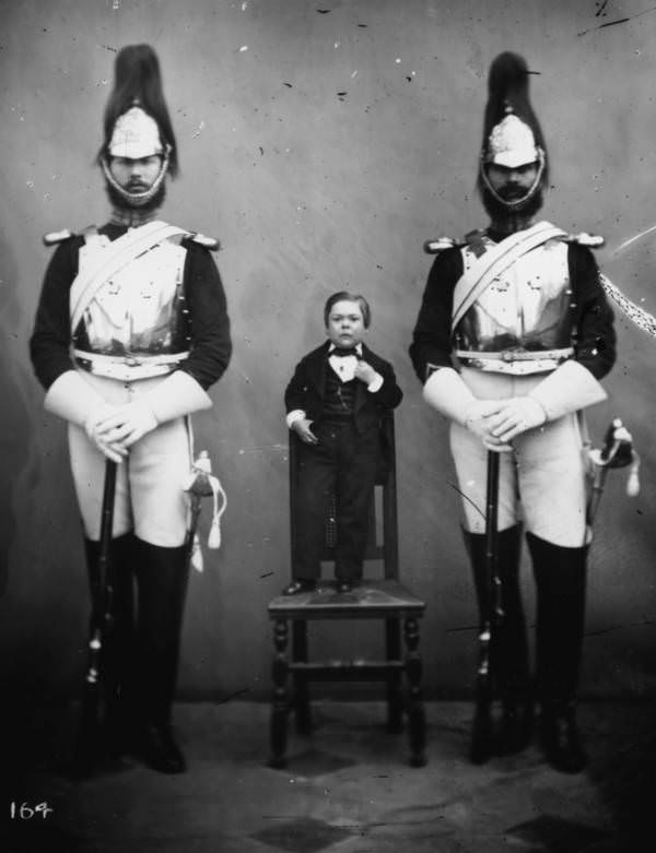 George Sherwood Stratton, better known as General Tom Thumb, standing on a chair between two guards, 1860