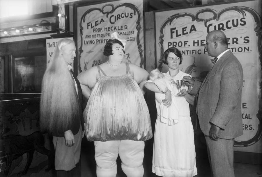At a Coney Island "freak" show an albino is photographed with the Fat Lady. A Flea Circus poster is in the background.
