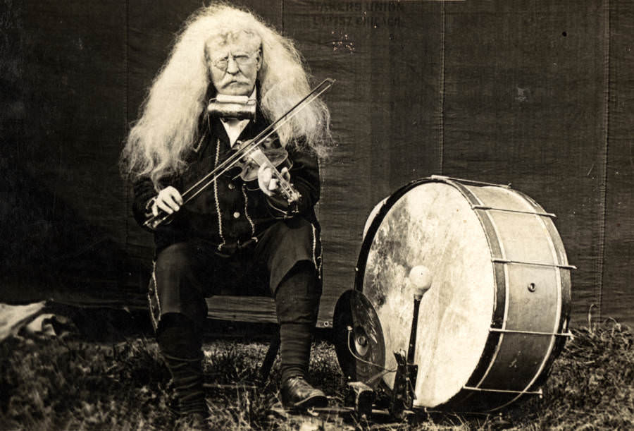 Elderly American musician "Professor" W.H. McMillan, a one-man band, sits with his bow and fiddle at the ready next to his drum and cymbal kit in front of a circus tent in Oakwood, Texas, 1910s