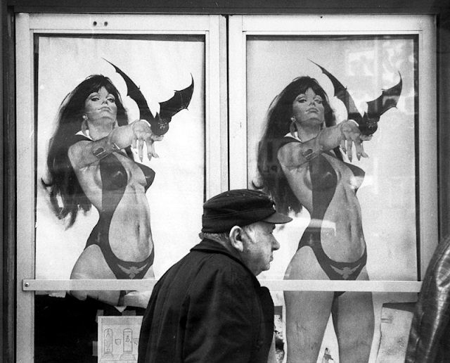 An elderly man walks past signs, posted on glass doors, with illustrations depicting a scantily clad woman holding a bat on her fingertips, New York, 1981.