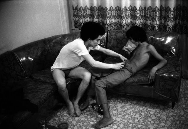 A 16-year-old child prostitute (right) sniffs glue out of a paper bag as his friend (left), an older hustler, undresses him in 1979.