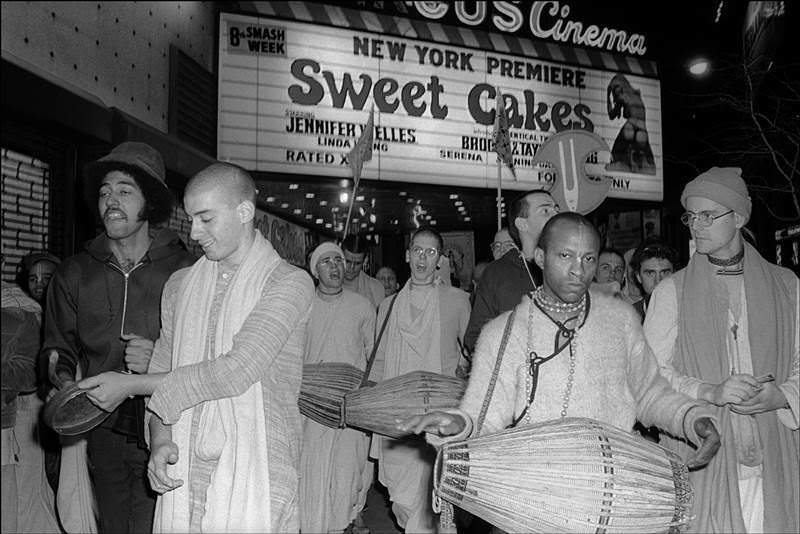 In 1976, a group of Hare Krishna followers sing and play instruments in Times Square under the marquee of an adult theater advertising the film Sweet Cakes.