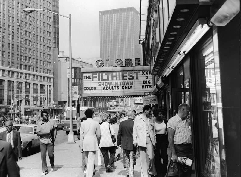 Tourists looking into the windows of Times Square as they pass under the marquee for the Globe theater advertising the "filthiest show in town.", 1975.