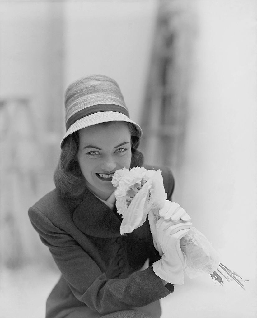 Ella Raines wearing hat by John-Frederics, holding flowers.