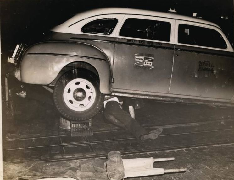 A police officer crouches under the rear end of a taxi jacked up on a crate and garbage can as the dead body of a man who was hit by the cab lies underneath, 1943