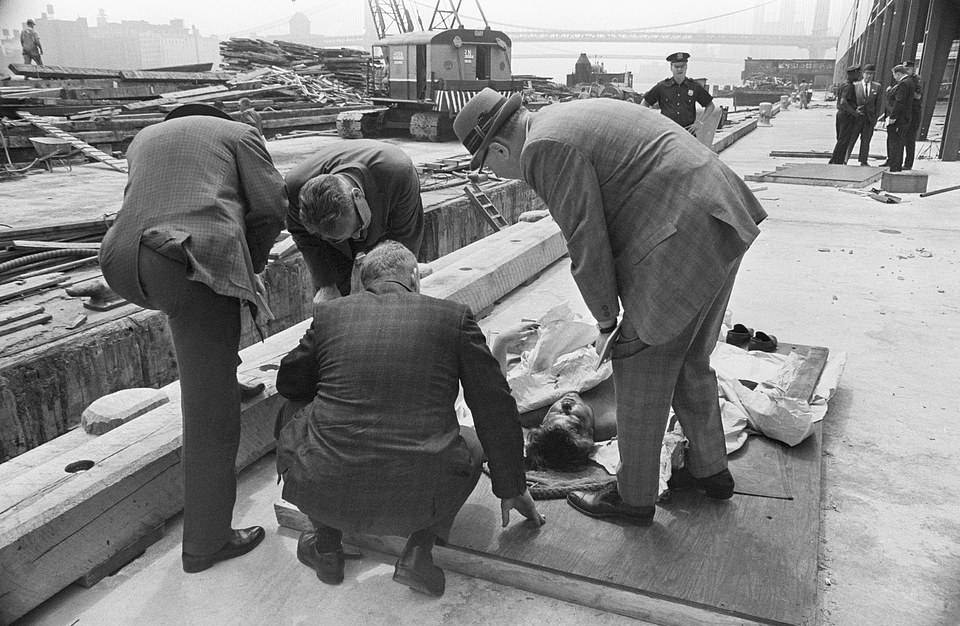 A medical examiner peering over the body of a man found floating in the East River in 1963. Bullet holes were found in the man's head
