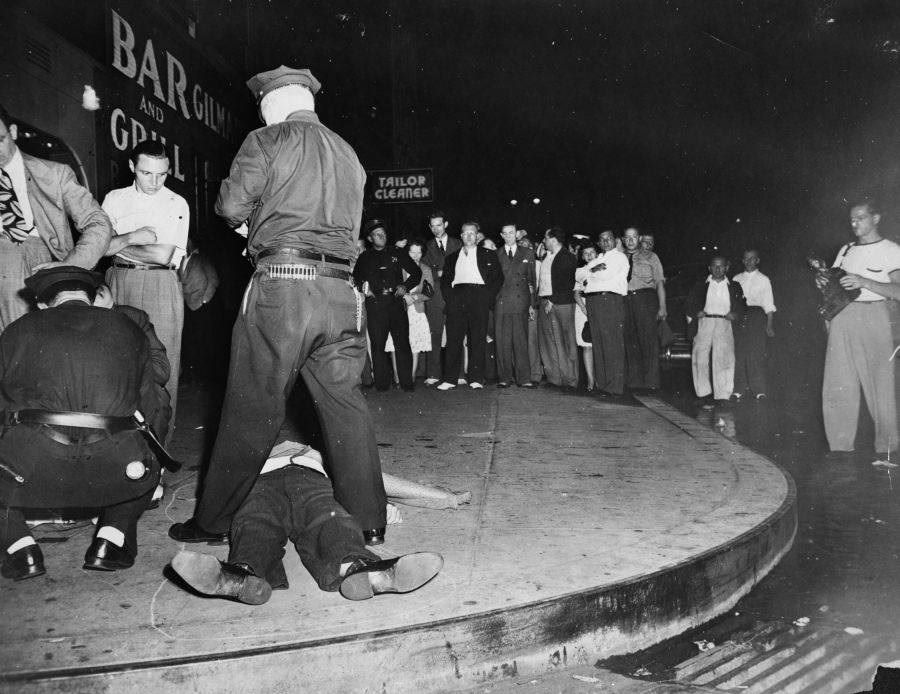 Watched by a curious crowd, a policeman straddles the body of a murder victim lying on the pavement outside a New York City bar, 1942.