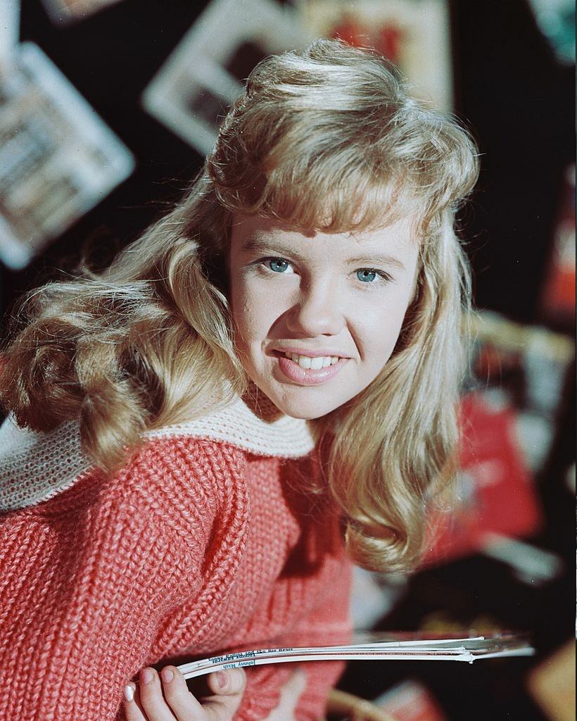 Hayley Mills wearing a coral pink-and-white knitted jumper and smiling while holding a number of LP record sleeves, 1960