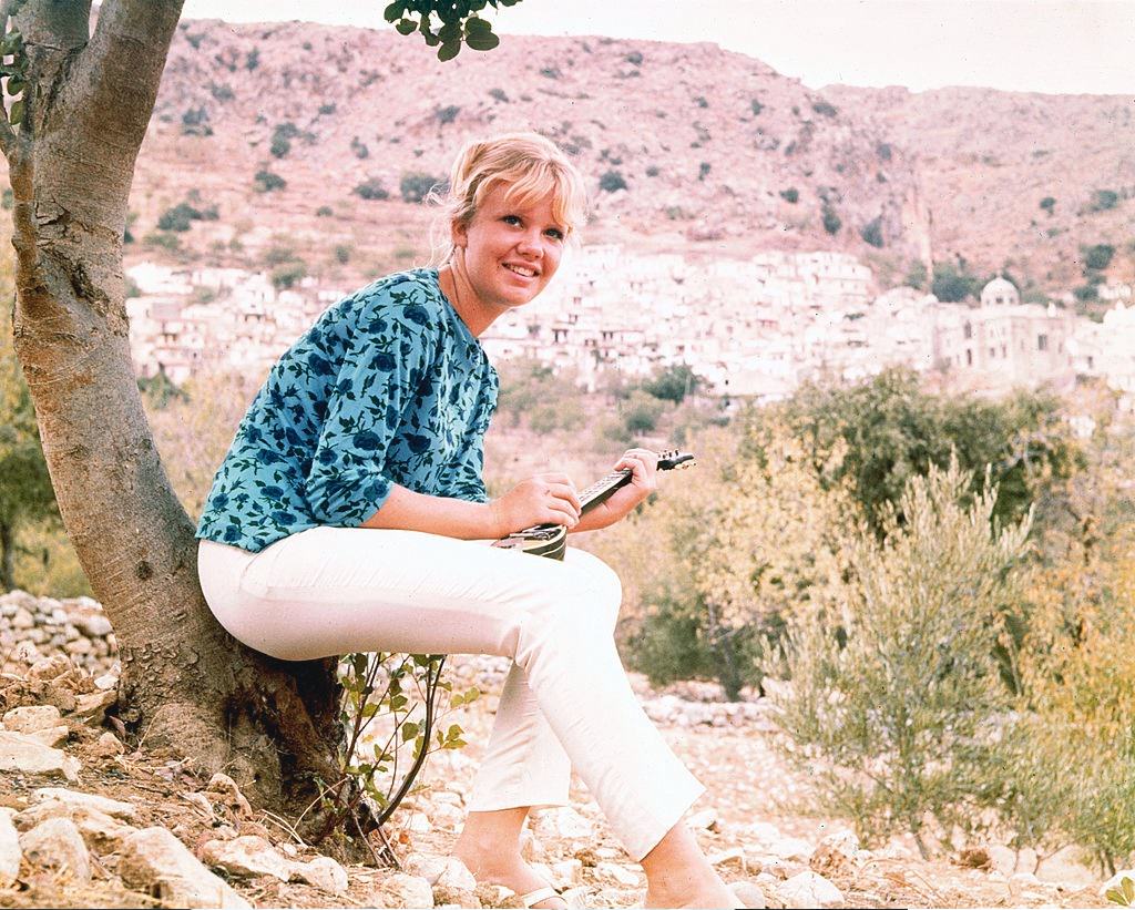 Hayley Mills wearing a blue blouse and white trousers and sitting at the base of a tree, 1965