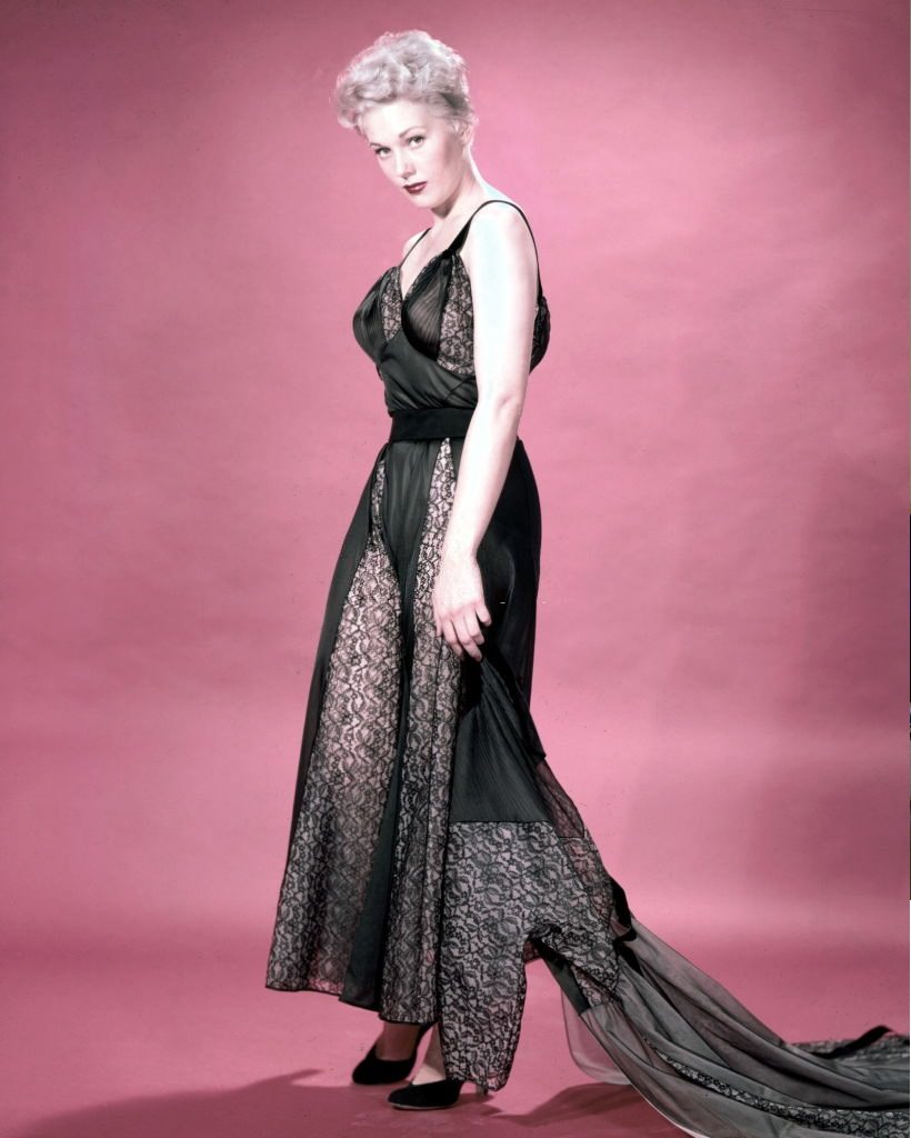 Kim Novak wearing a long black silk and black lace dress, with a black lace train, in a studio portrait, against a red background, circa 1960