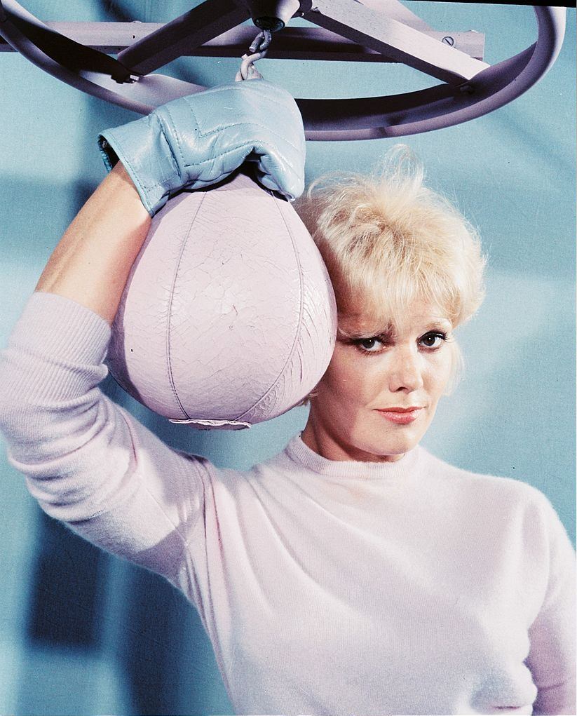 Kim Novak wearing a white woollen jumper and blue boxing gloves poses with a white punch ball, circa 1965