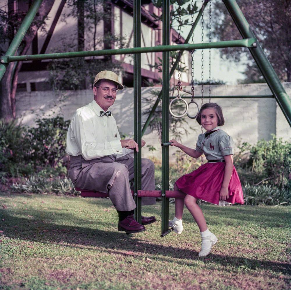 Comedian Groucho Marx sitting on a swingset with daughter Melinda, Beverly Hills, California, November 1953