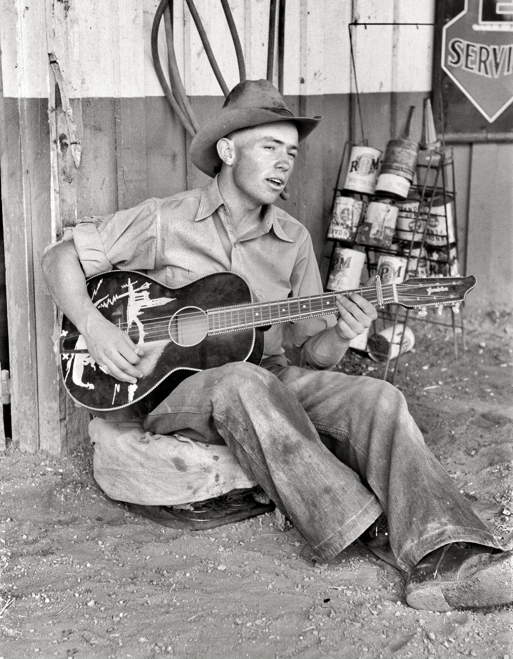 Farm boy playing guitar in front of the filling station and garage, Pie Town, New Mexico, June 1940