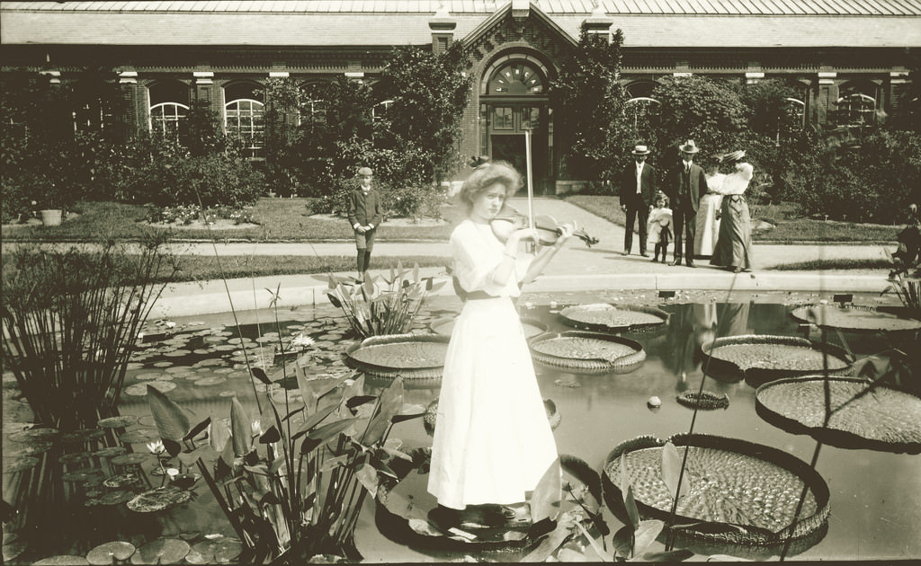 A young girl plays a violin while standing on lily pad in front of Linnean House at Shaw's Garden, 1900–1910.