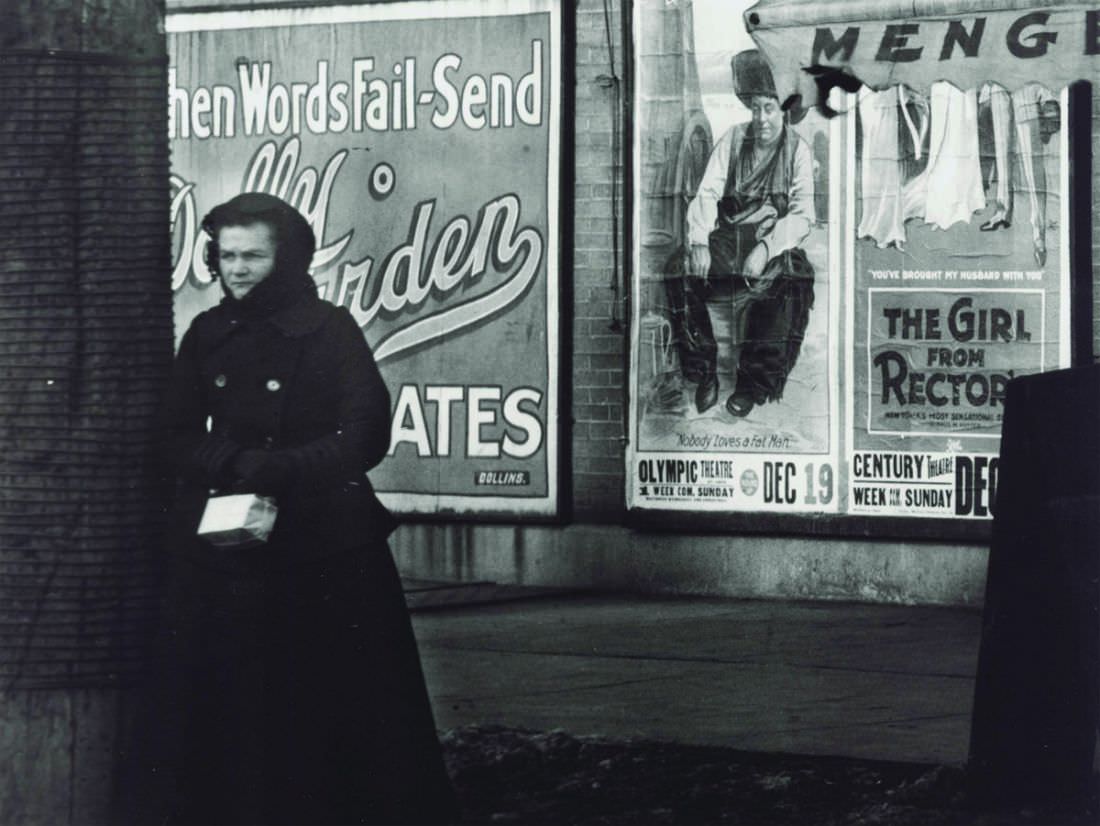 A woman bundled in winter wear at an unidentified street corner in front of advertisements for the movies The Round Up at the Olympic Theatre and The Girl from Rector’s showing at the Century Theatre, 1917