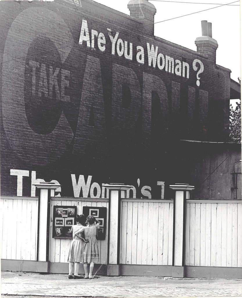 Two girls examining a bulletin board posted on a fence, ca. 1911–1921