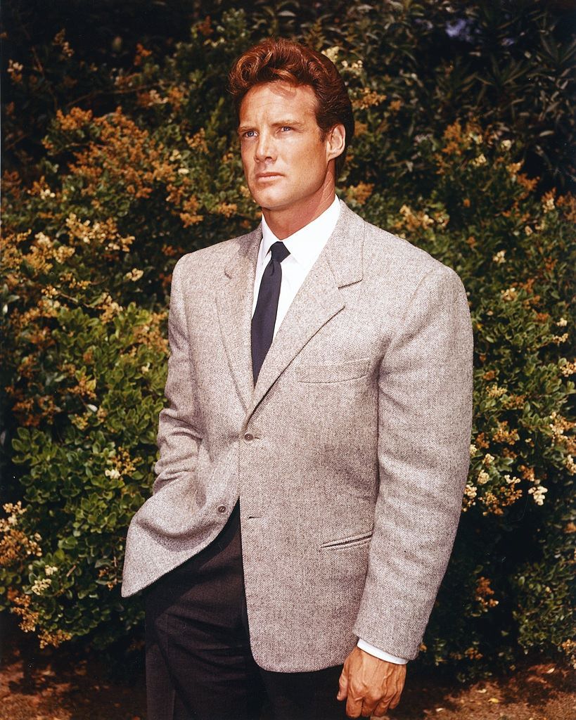Steve Reeves, wearing a grey jacket, white shirt and dark blue tie, posing in front of foliage, circa 1960