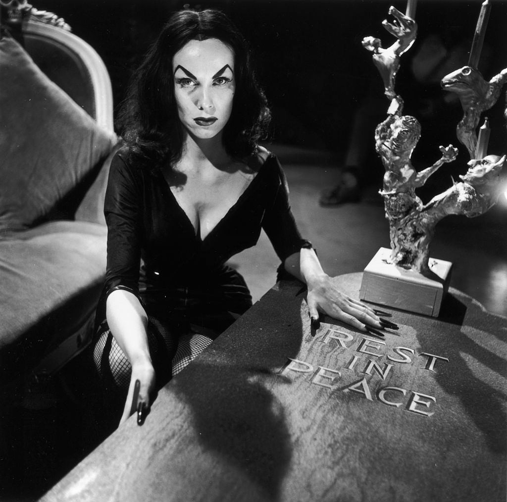 Maila Nurmi crouching beside a wooden coffin marked 'Rest in Peace'. She has arched, painted eyebrows and wears a low-cut black dress with fishnet stockings, 1956