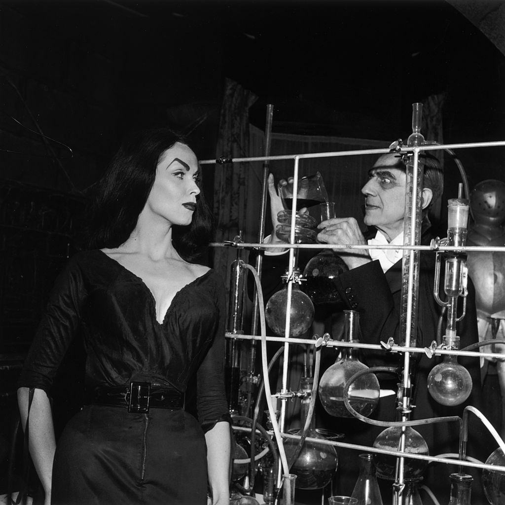 Bela Lugosi pouring a chemical into a test tube as Finnish-born actor and television host Vampira (Maila Nurmi) looks on beside an array of beakers and solutions, 1956