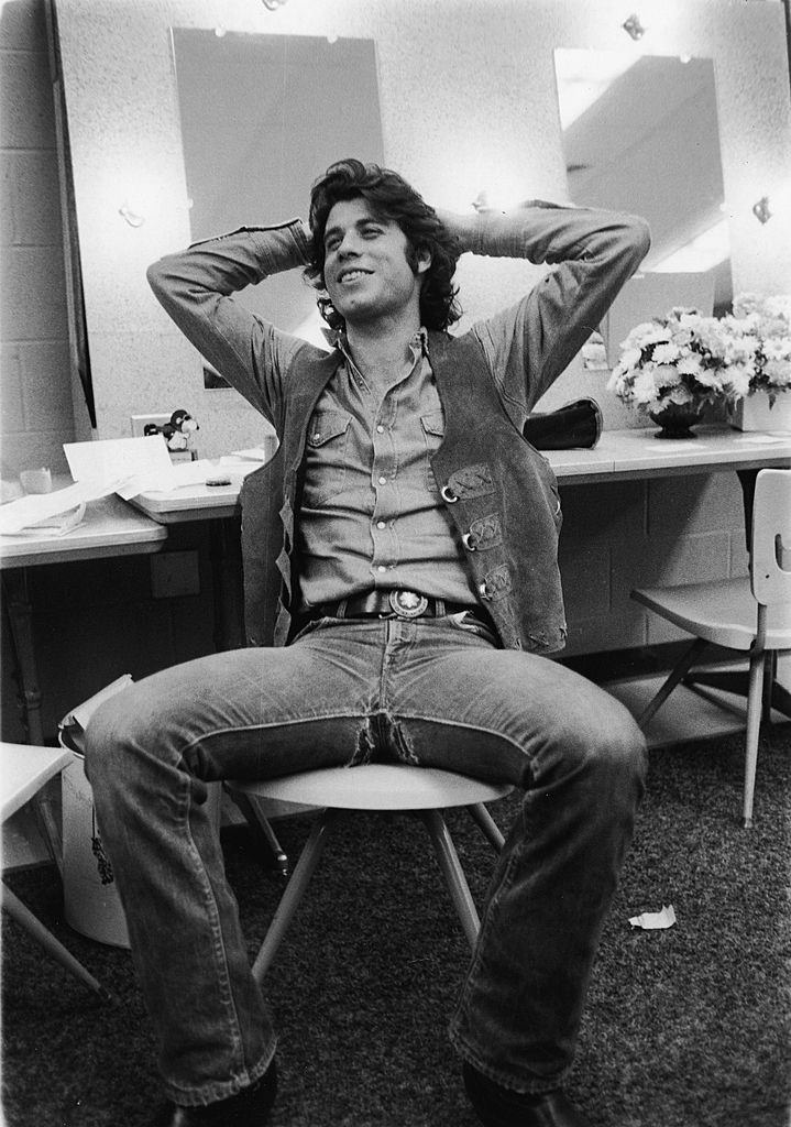 John Travolta sits in a chair and leans back with his hands behind his head, late 1970s